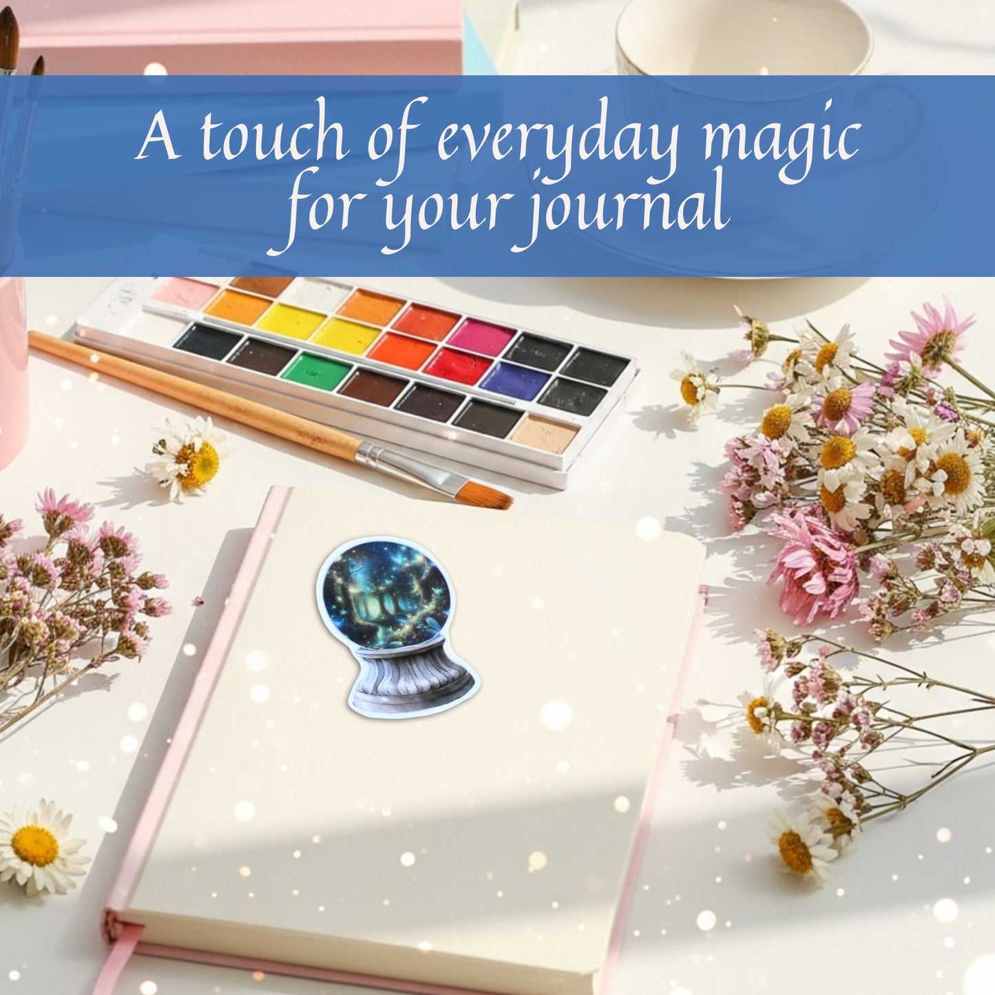 Journal with a whimsical forest night scene inside a crystal ball fantasy sticker, watercolor set, and flowers on a white surface with text 'A touch of everyday magic for your journal'.