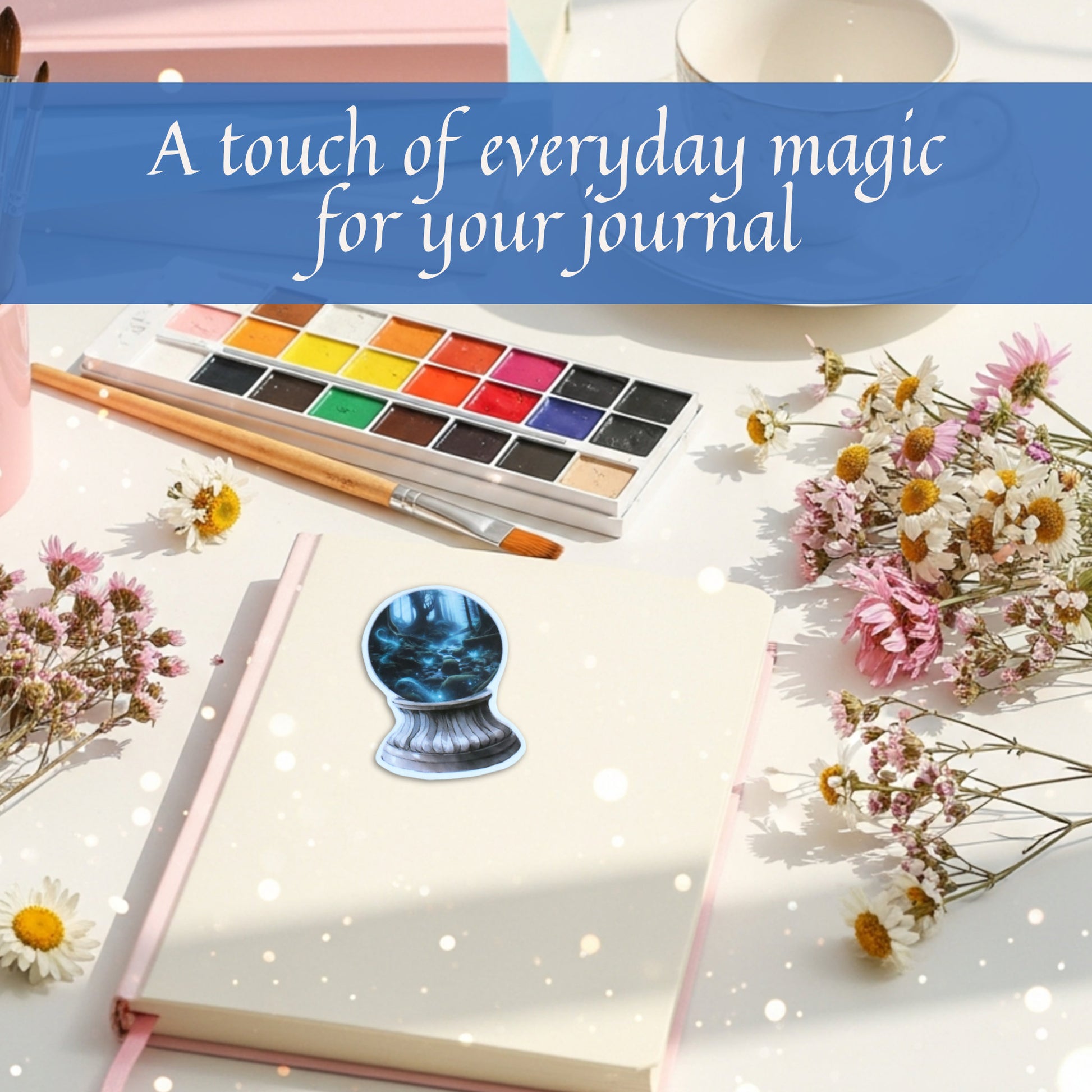 Journal with watercolor set and flowers on a white surface, with a fantasy landscape crystal ball sticker and text 'A touch of everyday magic for your journal'.